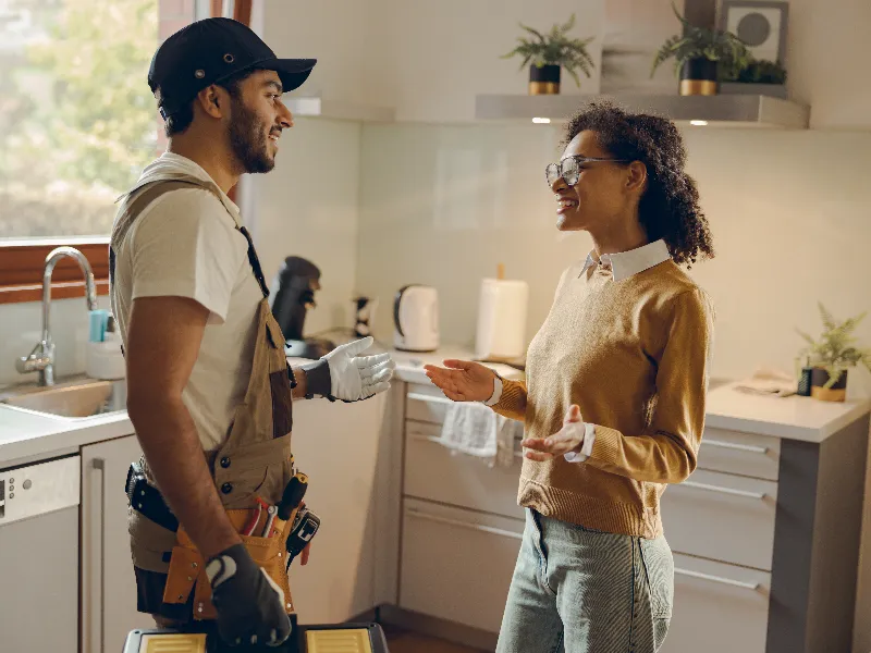 A repairman and a woman talk and smile in a kitchen.